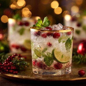 May include: A refreshing cocktail in a gold-rimmed glass, filled with ice, cranberries, lime slices, and mint leaves. The drink sits on a gold tray, with blurred Christmas lights in the background. A festive holiday beverage.