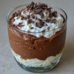 Op de afbeelding: Een gelaagd dessert in een helder glas. De onderste laag is een witte, romige substantie, gevolgd door een rijke, donkere chocolademousse. Het dessert is gegarneerd met slagroom en chocoladeschilfers. Het glas staat op een lichtgekleurd oppervlak.