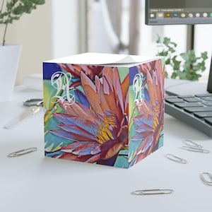 May include: A square notepad featuring a vibrant floral design with orange, purple, and yellow hues. The artwork depicts a close-up of a water lily with the letter "A" in white script. The notepad sits on a white surface.