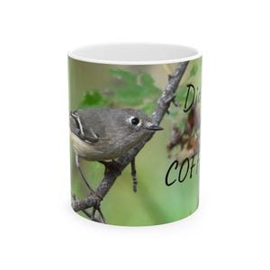 Puede incluir: Taza de café de cerámica blanca con una imagen detallada de un pájaro pequeño posado en una rama. El pájaro es gris y marrón con una raya blanca en su ala. La taza tiene las palabras "D COFFEE" en escritura negra.