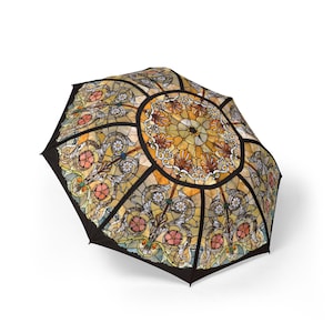 May include: An open umbrella with a stained-glass design. The umbrella features a circular pattern in the center with floral and geometric elements in shades of yellow, orange, and pink, outlined in black. The background is white.