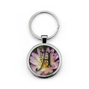 May include: A round silver keychain with a floral design. The keychain features a circular pendant with a pink and yellow flower background and a black letter "A" in the center. The keychain is made of metal.