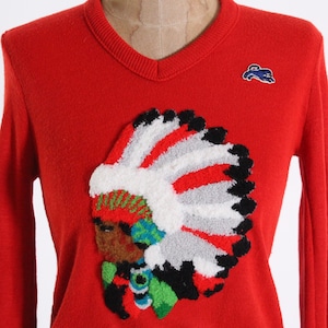 May include: Red knitted sweater with a large embroidered image of a Native American wearing a headdress. The headdress is made of white, red, and gray yarn and has black accents. The sweater has a V-neck and long sleeves.