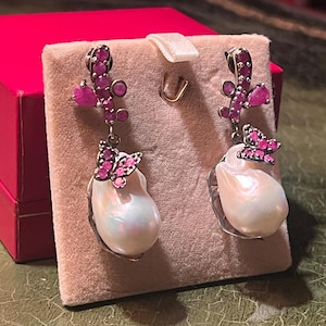 May include: A pair of dangle earrings featuring a large, irregular white pearl. The earrings have a silver-tone metal design with pink gemstone accents, including a butterfly shape. A pink box is in the background.