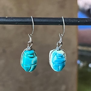 Op de afbeelding: Een paar turquoise scarabee-oorbellen. De oorbellen hangen aan zilverkleurige haken. De kevervormige kralen zijn fel turquoise van kleur met gesneden details. De oorbellen worden gepresenteerd tegen een vage achtergrond.