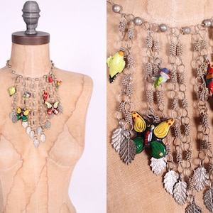 Puede incluir: Un collar de cadena plateada con múltiples dijes, incluyendo mariposas, pájaros y hojas. Los dijes están pintados en varios colores, incluyendo rojo, verde, amarillo y azul.