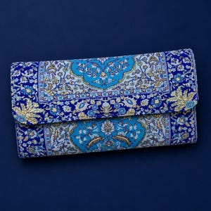 May include: A rectangular clutch bag featuring a vibrant blue, light blue, and white intricate floral pattern with metallic gold accents. The design mimics a traditional rug motif with a central medallion and ornate borders on a flap closure.