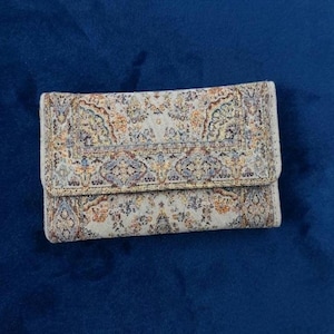 Op de afbeelding: Een rechthoekige portemonnee met een geweven stoffen buitenkant met een gedetailleerd, veelkleurig Perzisch tapijtpatroon in tinten beige, oranje, blauw en goud. De portemonnee heeft een overslagsluiting en een textuurachtige afwerking.