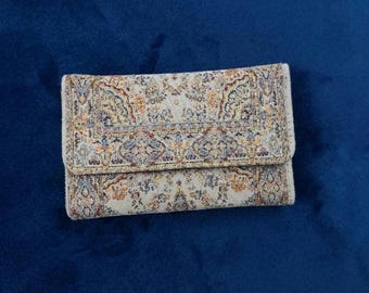 May include: A rectangular wallet featuring a woven fabric exterior with a detailed, multi-colored Persian-style rug pattern in shades of beige, orange, blue, and gold. The wallet has a fold-over flap closure and a textured, tapestry-like finish.