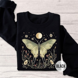 May include: Black crewneck sweatshirt featuring a large butterfly design with a moon and floral elements. The butterfly is light green with detailed wing patterns. The design includes a full moon, stars, and flowers. The word "BLACK" is printed on the sleeve.