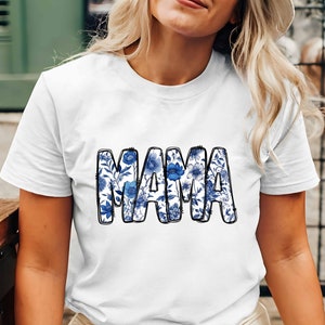 Könnte beinhalten: Weißes T-Shirt mit einem blauen Blumendesign, das "MAMA" buchstabiert.
