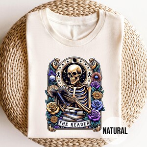 May include: A white t-shirt with a graphic of a skeleton reading a book. The graphic is surrounded by flowers and the text "The Reader" is below the skeleton. The text "NATURAL" is in the bottom right corner of the image.