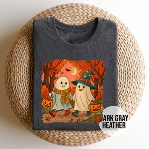 Könnte beinhalten: Dunkelgraues Heather-T-Shirt mit einem Halloween-Motiv. Die Grafik zeigt zwei Cartoon-Geister, die durch eine Herbstszene mit Kürbissen und Vollmond gehen. Der Text "ARK GRAY HEATHER" ist auf dem Shirt aufgedruckt.