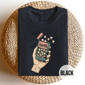 Könnte beinhalten: Schwarzes T-Shirt mit einer Grafik einer Hand, die eine Schachtel "teeth freshmints" hält. Die Schachtel ist geöffnet, wobei Zähne herausfallen. Fledermäuse sind im Hintergrund. Das Wort "teeth" ist auf der Schachtel aufgedruckt.