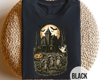 Puede incluir: Camiseta negra con un diseño de Halloween. El diseño incluye una casa embrujada, mapaches, un fantasma, una calabaza y murciélagos bajo la luna llena. El texto "BLACK" se muestra en la parte inferior derecha.