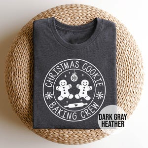 Könnte beinhalten: Dunkelgraues meliertes T-Shirt mit Rundhalsausschnitt. Das Shirt zeigt eine weiße kreisförmige Grafik mit dem Text "CHRISTMAS COOKIE BAKING CREW" um zwei Lebkuchenmänner und eine Teigrolle. Ein kleines Textfeld lautet "DARK GRAY HEATHER".