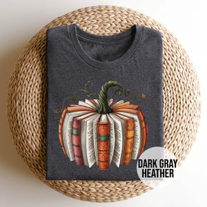May include: Dark gray heather t-shirt featuring a pumpkin design made of books. The pumpkin has an orange and brown color scheme with a green stem and vines. The text "DARK GRAY HEATHER" is in a white circle.