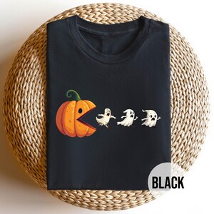May include: Black t-shirt featuring a Halloween-themed graphic. The design depicts an orange pumpkin with a wide open mouth chasing three white ghosts. The word "BLACK" is printed in a white circle at the bottom right.