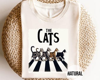 May include: Cream-colored t-shirt featuring a graphic of cats crossing a black and white striped crosswalk, with the words "THE CATS" above. The word "NATURAL" is printed below the shirt.