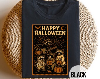 Puede incluir: Camiseta negra con un diseño temático de Halloween. El diseño incluye mapaches disfrazados, un sombrero de bruja, una calabaza y el texto "HAPPY HALLOWEEN". La camiseta se muestra sobre una superficie circular tejida.