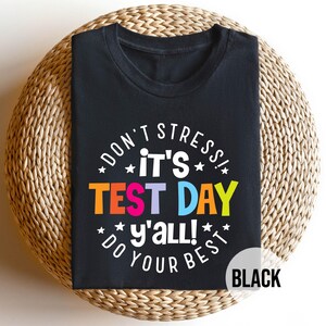 May include: A black short-sleeved t-shirt featuring white text that reads "DON'T STRESS! IT'S TEST DAY Y'ALL! DO YOUR BEST" with the words "TEST DAY" in orange, purple, blue, and yellow, accented by small white stars in a circular arrangement.