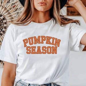 Könnte beinhalten: Weißes T-Shirt mit dem orangefarbenen Text "PUMPKIN SEASON" in einer Schriftart im College-Stil. Das T-Shirt hat einen klassischen Rundhalsausschnitt. Der Text ist mittig auf der Vorderseite platziert.