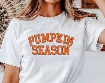 Puede incluir: Camiseta blanca con el texto naranja "PUMPKIN SEASON" en una fuente de estilo universitario. La camiseta es de estilo cuello redondo clásico. El texto está centrado en la parte delantera.