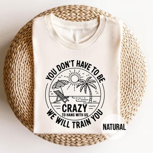 May include: A folded, natural-colored t-shirt with a circular graphic featuring a beach scene with a palm tree, sun, and text that reads "YOU DON'T HAVE TO BE CRAZY TO HANG WITH US WE WILL TRAIN YOU". The word "NATURAL" is printed below the graphic.