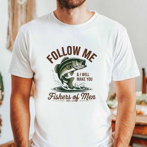 Könnte beinhalten: Weißes T-Shirt mit einer Grafik eines springenden Fisches und dem Text "FOLLOW ME & I WILL MAKE YOU Fishers of Men". Das Design zeigt einen grün-schwarzen Fisch mit Wasserspritzern in brauner Schrift.