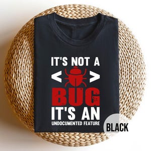 May include: Black t-shirt with the text "IT'S NOT A BUG IT'S AN UNDOCUMENTED FEATURE" in white and red. The design includes a red bug graphic and code brackets. The shirt is displayed on a woven, circular surface.