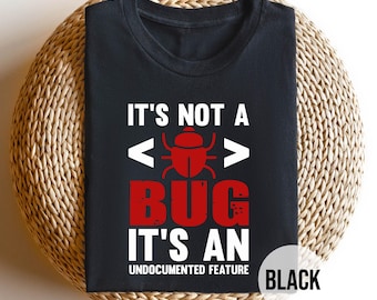 May include: Black t-shirt with the text "IT'S NOT A BUG IT'S AN UNDOCUMENTED FEATURE" in white and red. The design includes a red bug graphic and code brackets. The shirt is displayed on a woven, circular surface.