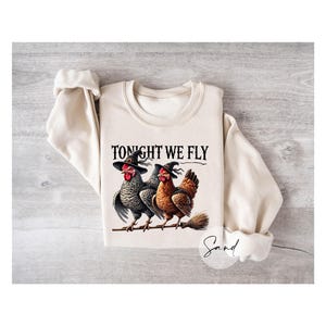 May include: Beige sweatshirt with the text "TONIGHT WE FLY". The graphic features two chickens wearing witch hats, perched on a branch with a broom. The chickens are black and white, and brown.