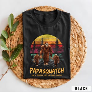 May include: Black t-shirt featuring a Bigfoot family graphic with a sunset backdrop. The design includes the text "Papasquatch" and "Like a Grandpa, Just Way More Squatchy". The shirt is displayed on a woven mat.
