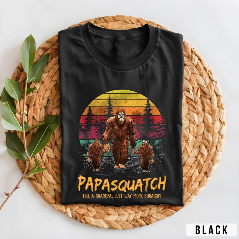 May include: Black t-shirt featuring a Bigfoot family graphic with a sunset backdrop. The design includes the text "Papasquatch" and "Like a Grandpa, Just Way More Squatchy". The shirt is displayed on a woven mat.