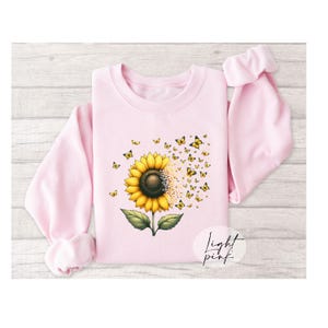 K&ouml;nnte beinhalten: Ein hellrosa Sweatshirt mit einem Sonnenblumen-Motiv. Die Sonnenblume hat gelbe Bl&uuml;tenbl&auml;tter und eine braune Mitte. Schmetterlinge fliegen aus der Sonnenblume. Der Text "Light Pink" ist in der rechten unteren Ecke des Sweatshirts gedruckt.