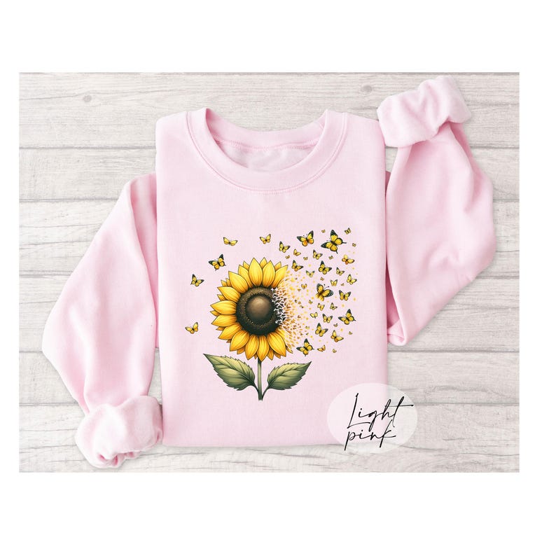 K&ouml;nnte beinhalten: Ein hellrosa Sweatshirt mit einem Sonnenblumen-Motiv. Die Sonnenblume hat gelbe Bl&uuml;tenbl&auml;tter und eine braune Mitte. Schmetterlinge fliegen aus der Sonnenblume. Der Text "Light Pink" ist in der rechten unteren Ecke des Sweatshirts gedruckt.