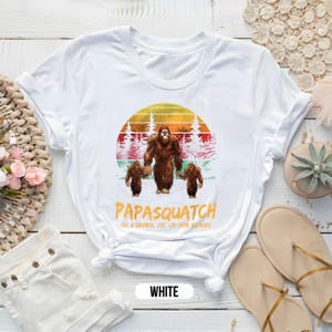 May include: White t-shirt featuring a Bigfoot family graphic. The design includes a large Bigfoot holding hands with two smaller ones, set against a sunset backdrop. The text "Papasquatch" and "Like a Grandpa, Just Way More Squatchy" are printed below.