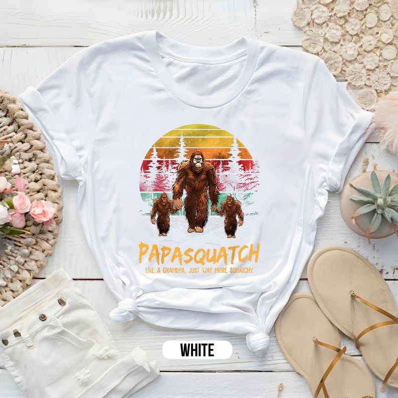 May include: White t-shirt featuring a Bigfoot family graphic. The design includes a large Bigfoot holding hands with two smaller ones, set against a sunset backdrop. The text "Papasquatch" and "Like a Grandpa, Just Way More Squatchy" are printed below.
