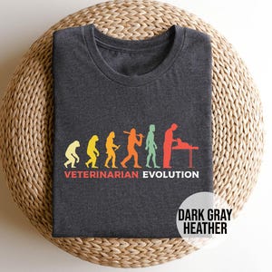 May include: Dark gray heather t-shirt with a graphic of the evolution of a veterinarian, starting with a caveman and ending with a veterinarian examining a pet. The text "Veterinarian Evolution" is printed below the graphic.