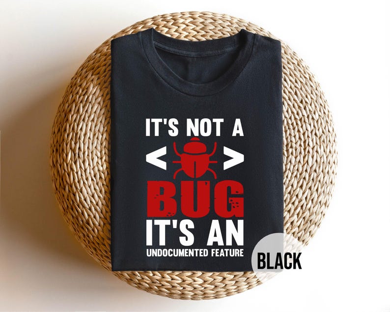 May include: Black t-shirt with the text "IT'S NOT A BUG IT'S AN UNDOCUMENTED FEATURE" in white and red. The design includes a red bug graphic and code brackets. The shirt is displayed on a woven, circular surface.