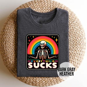 Pu&ograve; includere: T-shirt in grigio scuro melange con una grafica di arcobaleno e scheletro. Il testo "Everything Sucks" &egrave; stampato in un font colorato.