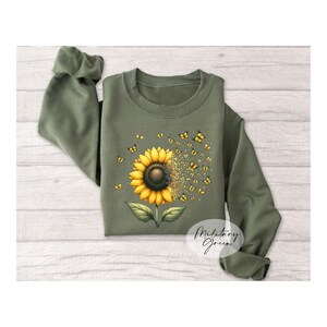 K&ouml;nnte beinhalten: Ein gr&uuml;nes Sweatshirt mit einem Sonnenblumen-Motiv. Die Sonnenblume besteht aus gelben Bl&uuml;tenbl&auml;ttern und einer braunen Mitte. Schmetterlinge fliegen aus der Sonnenblume. Der Text "Military Green" steht in der rechten unteren Ecke des Sweatshirts.