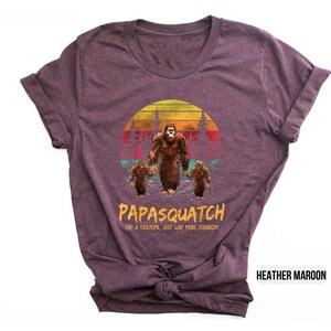 May include: Heather maroon t-shirt featuring a Bigfoot family graphic. The design includes a large Bigfoot, two smaller ones, and the text "PAPASQUATCH like a grandpa just way more squatchy" in yellow and orange.
