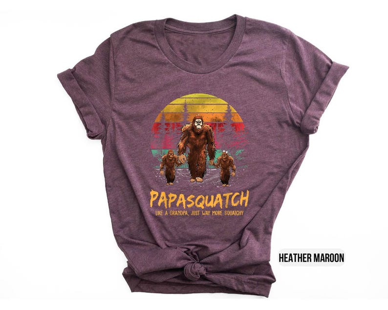 May include: Heather maroon t-shirt featuring a Bigfoot family graphic. The design includes a large Bigfoot, two smaller ones, and the text "PAPASQUATCH like a grandpa just way more squatchy" in yellow and orange.