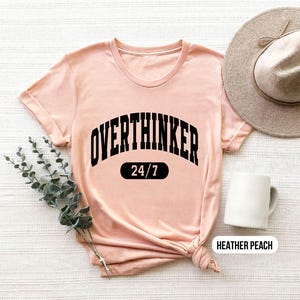 Peut inclure: Un t-shirt rose p&acirc;le avec un texte noir qui dit "Overthinker 24/7". Le t-shirt est fait d'un tissu doux et confortable et a une coupe d&eacute;contract&eacute;e.