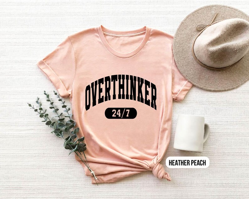 Peut inclure: Un t-shirt rose p&acirc;le avec un texte noir qui dit "Overthinker 24/7". Le t-shirt est fait d'un tissu doux et confortable et a une coupe d&eacute;contract&eacute;e.