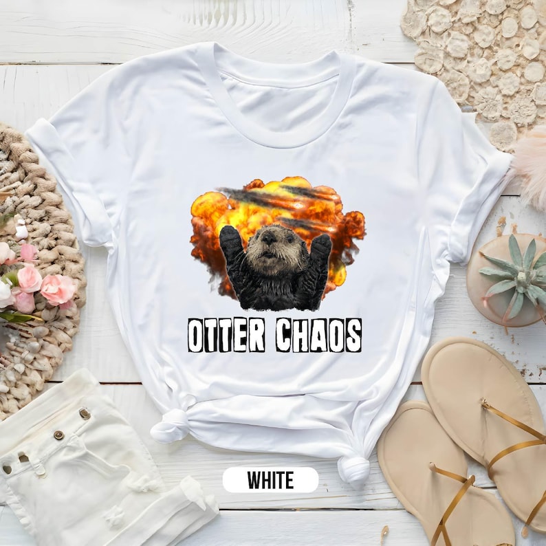 May include: White t-shirt with a graphic of an otter with its arms raised in the air, surrounded by an explosion. The text "Otter Chaos" is printed below the graphic.