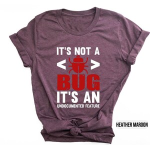 May include: Heather maroon t-shirt with the text "IT'S NOT A BUG IT'S AN UNDOCUMENTED FEATURE" in white and red. The design includes a bug graphic. The shirt is a casual wear item.