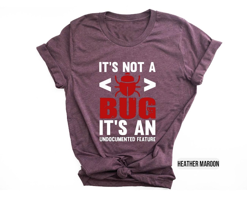 May include: Heather maroon t-shirt with the text "IT'S NOT A BUG IT'S AN UNDOCUMENTED FEATURE" in white and red. The design includes a bug graphic. The shirt is a casual wear item.