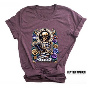 May include: A heather maroon t-shirt with a graphic of a skeleton reading a book surrounded by flowers. The text "The Reader" is printed below the skeleton.